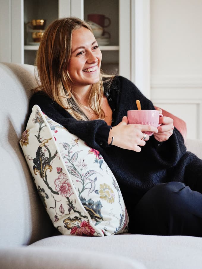 Frau mit einer rosa Kaffeetasse auf einem grauen Sofa mit geblümtem Kissen.