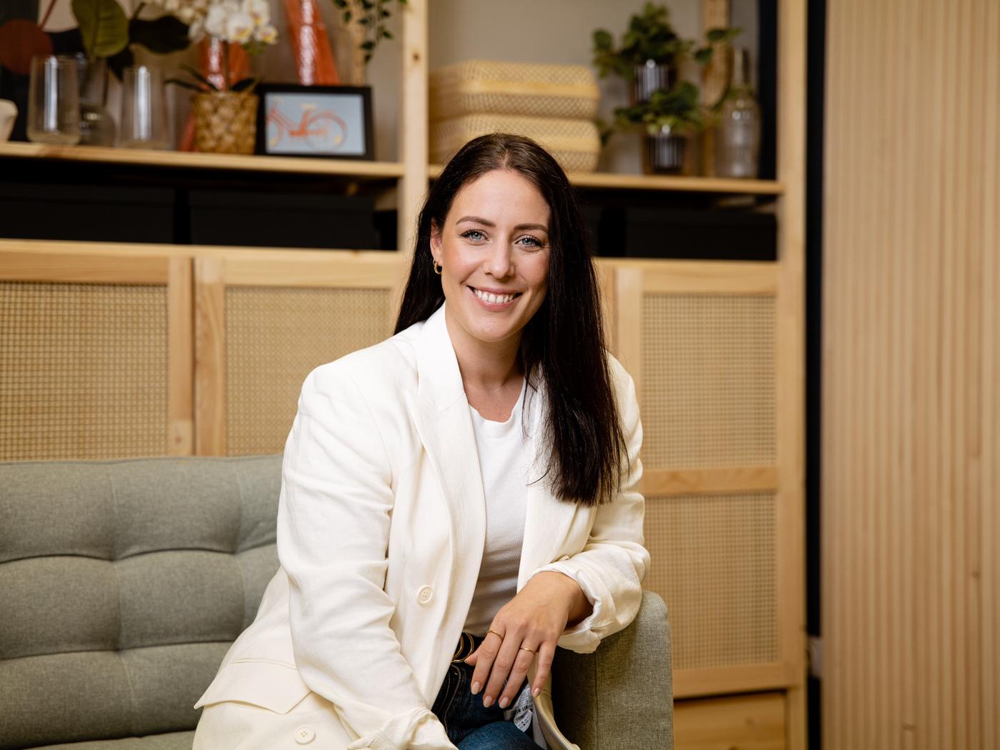 Frau mit weißem Blazer sitzt lächelnd auf einem Sofa in modernem IKEA-Einrichtungsstil mit Holz- und Naturmaterialien im Hintergrund.