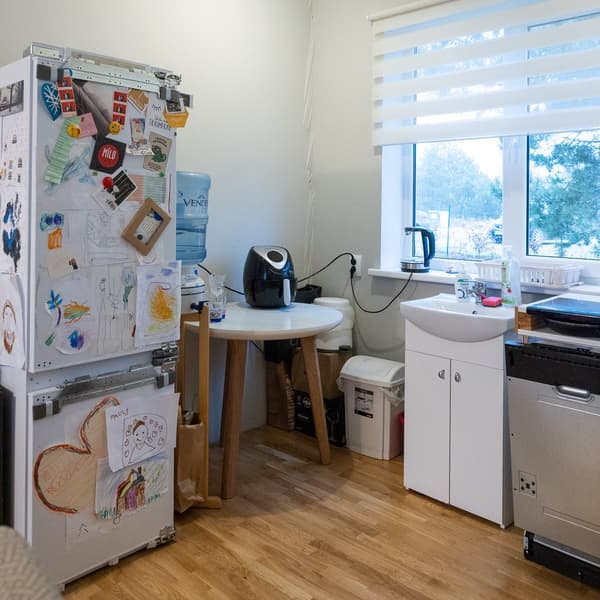 Fridge covered with magnets next to a workspace with a table and chairs; everyday household items visible on the table and shelves.