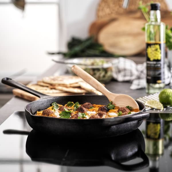 Frigideira em preto com legumes e uma colher em madeira, sobre uma placa.