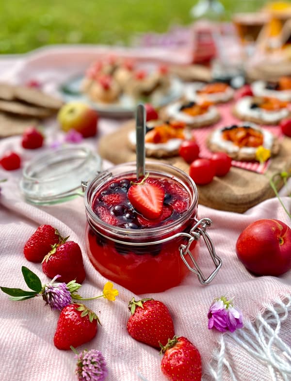 Frische Erdbeermarmelade in einem Glasgefäss mit einem Löffel, umgeben von ganzen Erdbeeren und einer Picknickkulisse im Hintergrund.