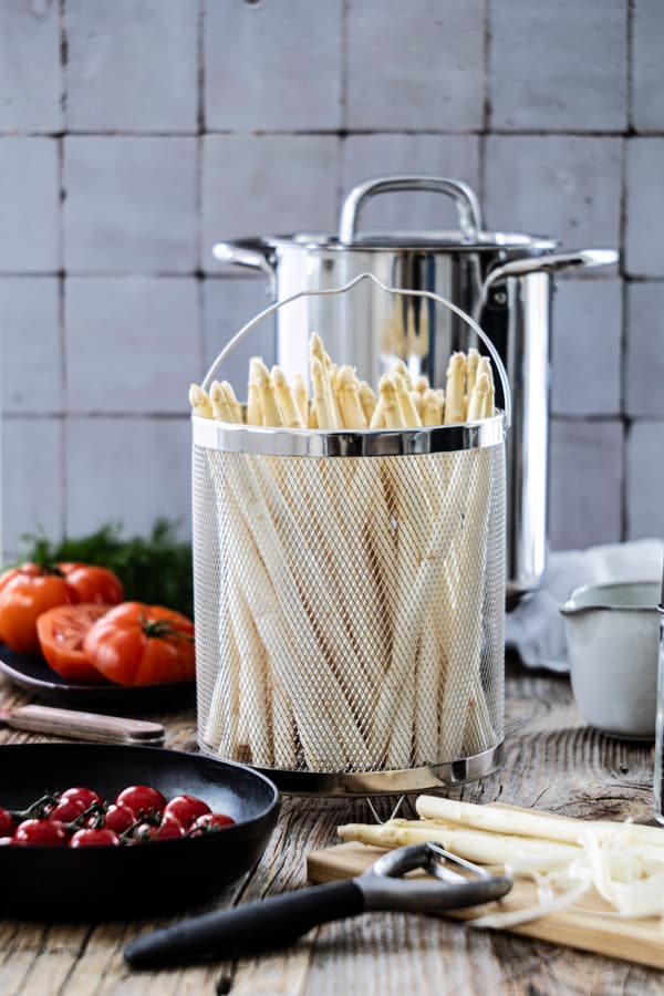 Frischer Spargel steht in einem Garkorb vor einem Kochtopf.