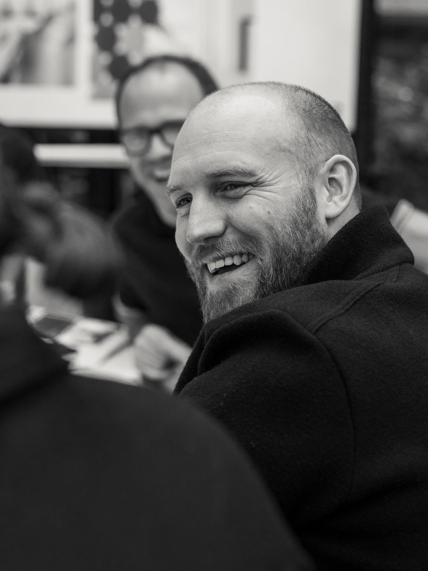 Friso Wiersma IKEA-ko diseinatzailea, jaka beltzarekin, norbaiti begira dago irribarrez; mahai batean eserita dago, irribarrez dauden beste pertsona batzuen ondoan.