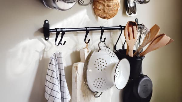 From a black HULTARP rail hang a tea towel, a cutting board, a white colander and utensils in a black HULTARP container.