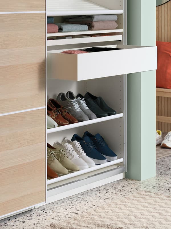 From a PAX wardrobe extends a white KOMPLEMENT pull-out tray with shoe insert and four pairs of different coloured sneakers.