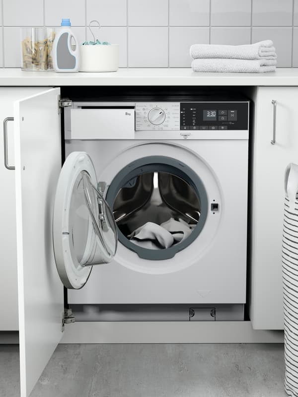 Frontlader-Waschmaschine unter einer Arbeitsplatte in einem Hauswirtschaftsraum oder einer Küche; die Tür der Maschine ist geöffnet und zeigt Kleidung im Inneren. Auf der Arbeitsplatte darüber liegen gefaltete Handtücher, eine Flasche Waschmittel und weitere Reinigungsutensilien.