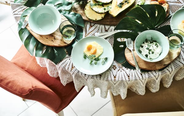 Fruits et yaourts servis sur une table décorée de grandes feuilles, de planches en bois et de vaisselle verte.