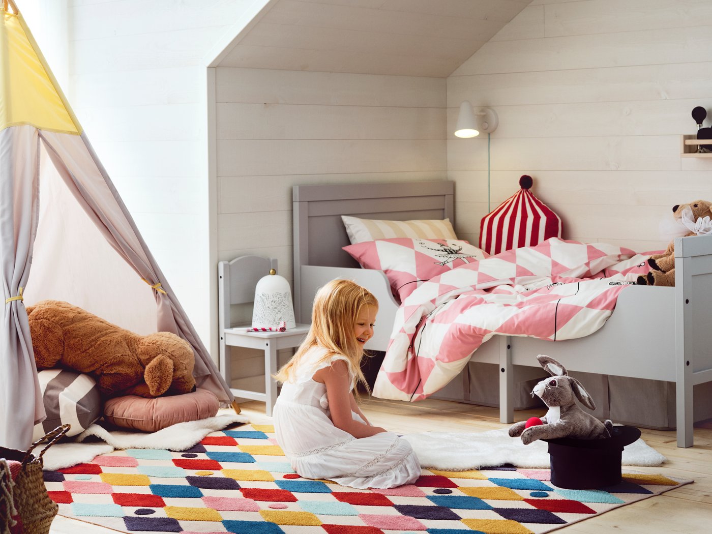 فتاة تجلس على سجادة BUSENKEL ألوانها زاهية في غرفة نوم بالقرب من سرير قابل للتمديد SUNDVIK رمادي وتنظر إلى دمية على شكل حيوان.