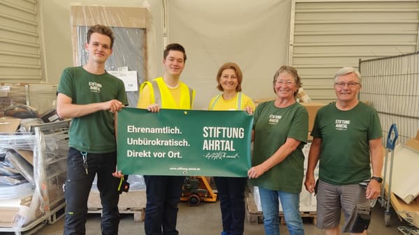 Fünf Personen, darunter zwei IKEA-Mitarbeitende in gelben Warnwesten und drei Mitglieder der Stiftung Ahrtal in grünen T-Shirts, halten gemeinsam ein grünes Banner mit der Aufschrift „Ehrenamtlich. Unbürokratisch. Direkt vor Ort. Stiftung Ahrtal“. Im Hintergrund sind Paletten mit Möbeln und Kartons gestapelt.  