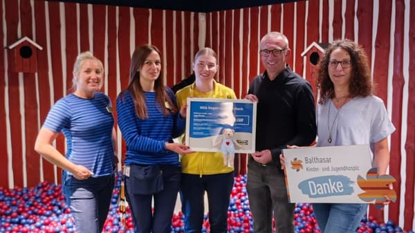 Fünf Personen stehen in einem IKEA-Kinderbereich mit roten Holzwänden und einem Bällebad. Drei von ihnen tragen IKEA-Mitarbeiterkleidung und überreichen einen Spendenscheck über 3.500 Euro an Vertreter:innen des Balthasar Kinder- und Jugendhospizes, das mit einem „Danke“-Schild darauf hinweist.  