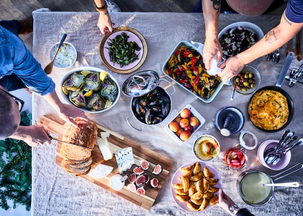 Full table family dinner with VARDAGEN plates and oven dish and a person cutting som bread on SMÅÄTA chopping board.