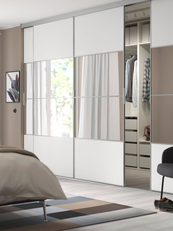 Full view of a SKYTTA wardrobe with sliding doors in MEHAMN white and AULI mirror panels. The wardrobe is slightly open, to reveal clothes neatly hung above drawers.