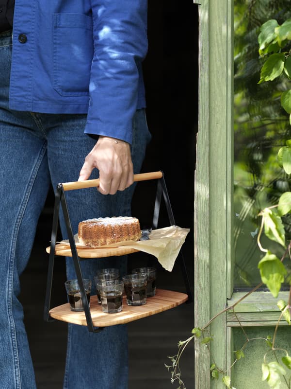 FULLSPÄCKAD serving tray made in bamboo with two tiers holding cake and drinks is being carried by its handle.