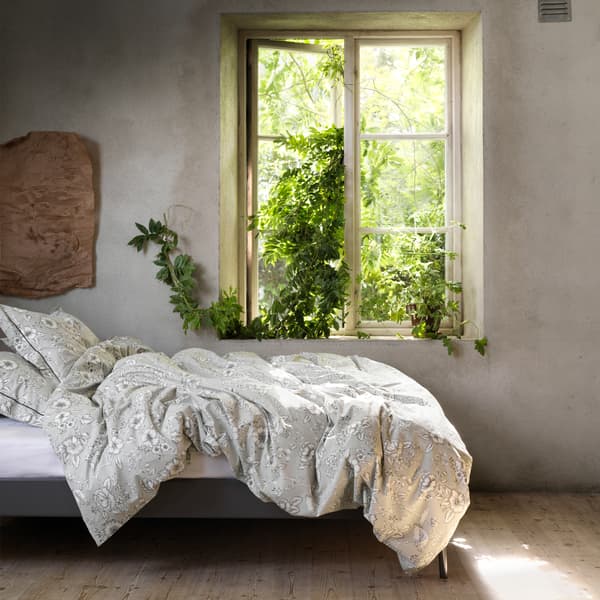 Funda de edredón e almofada PRAKTBRÄCKA con deseño de flores tradicional nunha cómoda cama xunto a unha ventá aberta.