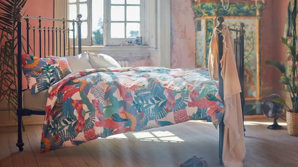 Funda nórdica SKOGSFIBBLA con un estampado exótico aclarado por el sol sobre la cama de una amplia habitación con grandes ventanas.