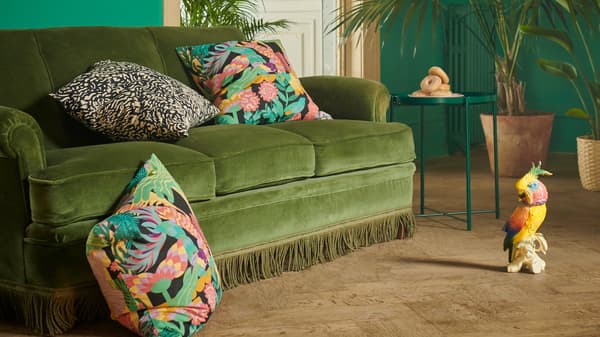 Fundas para coxíns NÄBBFLY e GRIMHILD con deseños exóticos e coloridos sobre un sofá de veludo verde dentro dunha habitación rodeada de plantas.
