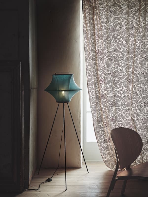 FYXNÄS floor lamp with a spacey design in turquoise knitted fabric and metal tripod legs standing in a corner near a window.