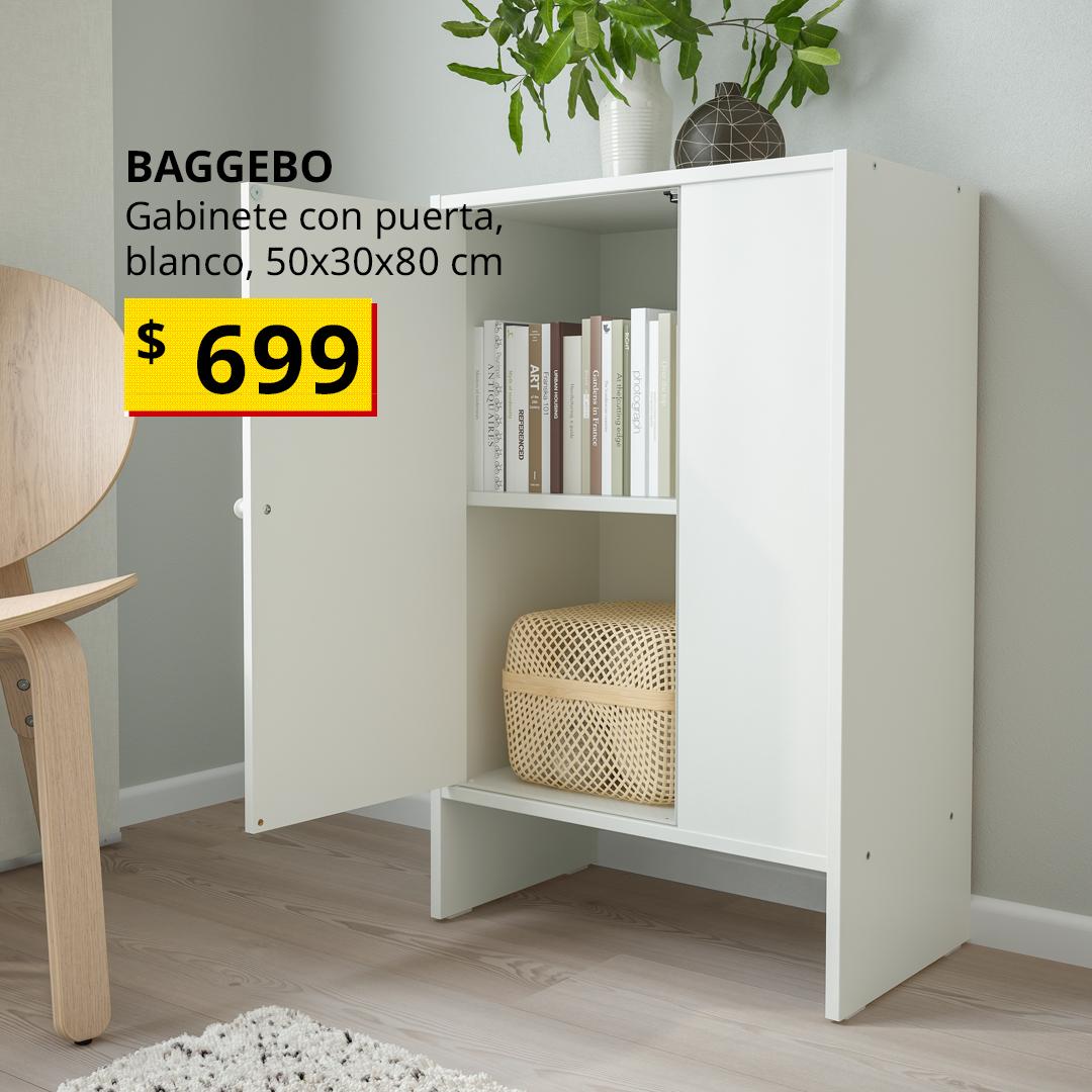 Gabinete BAGGEBO blanco con puerta en sala minimalista, estante superior con libros y canasta de almacenamiento, decoración con jarrón de ramas verdes y florero oscuro geométrico; diseño compacto, funcional y elegante para interiores modernos.