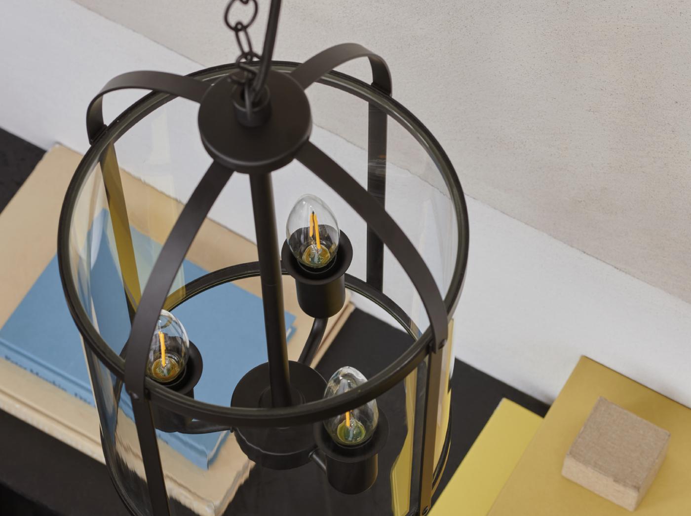 ภาพระยะใกล้ของโคมแขวนเพดาน GALJON ดีไซน์รูปตะเกียงที่มีหลอดไฟประดับสามดวงแขวนอยู่ และมีหนังสือสีสดใสเป็นพื้นหลัง