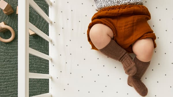 Gambe e parte del busto di un bambino con calze e pantaloncini marroni steso su un lenzuolo con angoli LENAST a pois all'interno di un lettino bianco - IKEA.