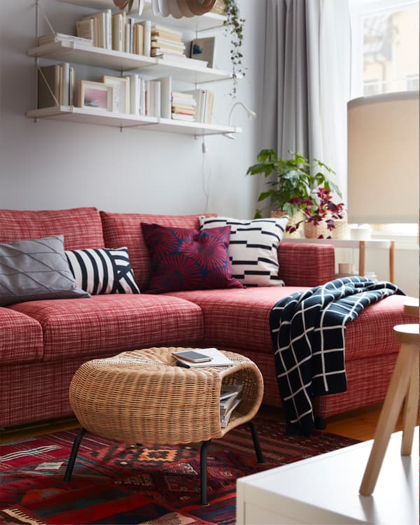 GAMLEHULT rattan footstool with storage on a red patterned rug, a red sofa, white wall-mounted shelves and a checkered throw.
