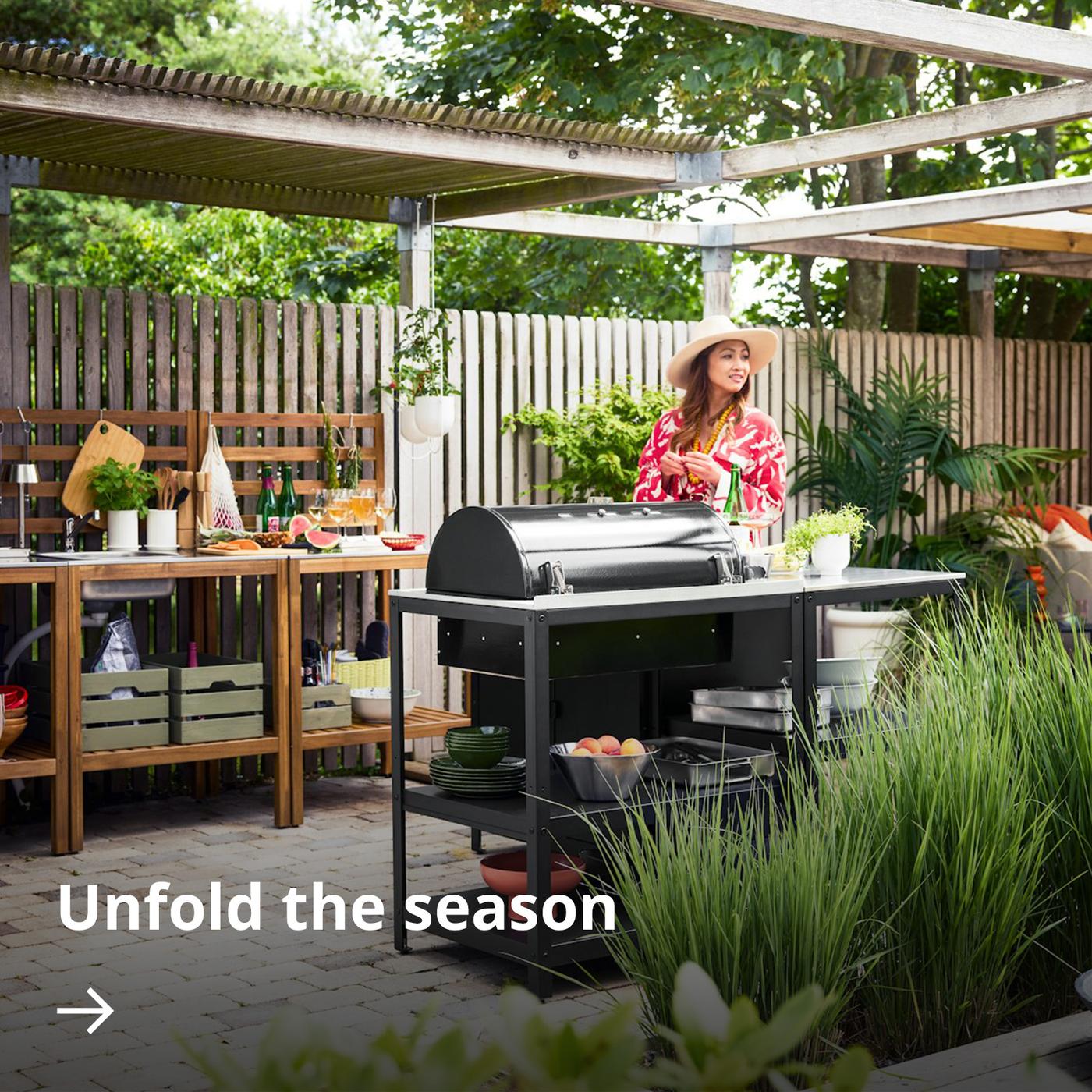 Garden scene with a black GRILLSKÄR charcoal barbecue and wooden NÄMMARÖ outdoor kitchen units under a pergola.