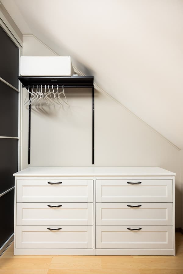 Garderobes iekšskats ar atvilktnēm, plauktiem un drēbju stangām, kas pielāgotas slīpiem griestiem efektīvai uzglabāšanai.