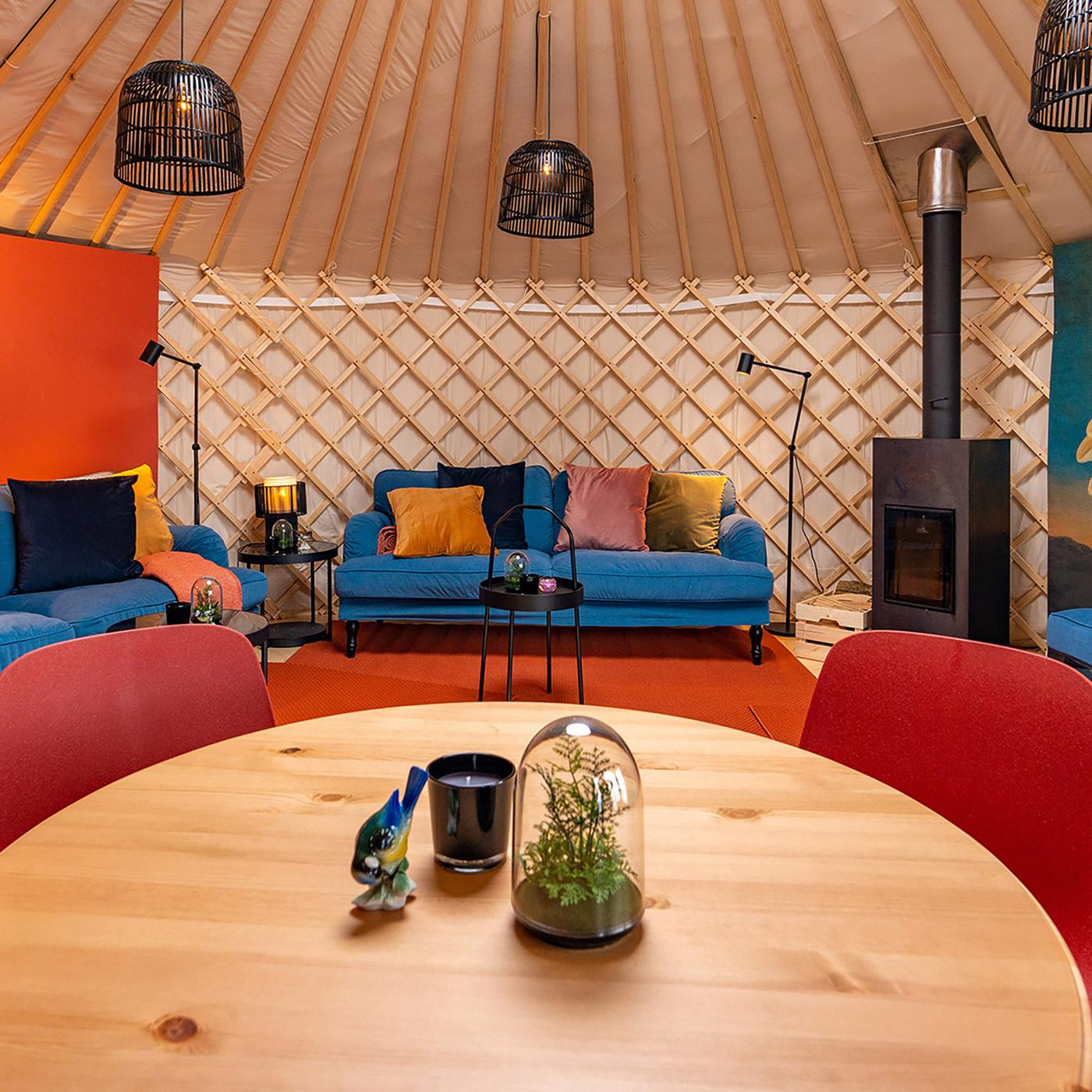Gemütliche Jurte mit blauen Sofas, Holztisch, Pflanzen, Ofen, Lampen und oranger Wand.