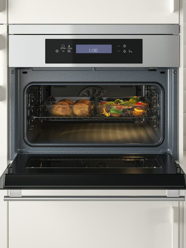 Geöffneter Backofen mit digitalem Bedienfeld oben; im beleuchteten Innenraum befindet sich ein Blech mit mehreren Lebensmitteln, die gerade gegart werden.