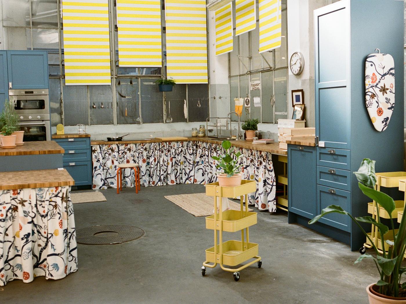 Gezeigt wird eine große Küche mit blaue Fronten, bunten Vorhängen, gelbes Servierwagerl im Fokus