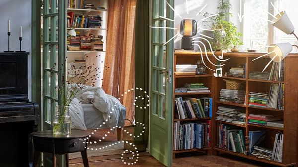 Gezellige woonkamer met houten vloer, zwarte piano en boekenkast, met zicht op een lichtrijke slaapkamer via groene openslaande deuren.