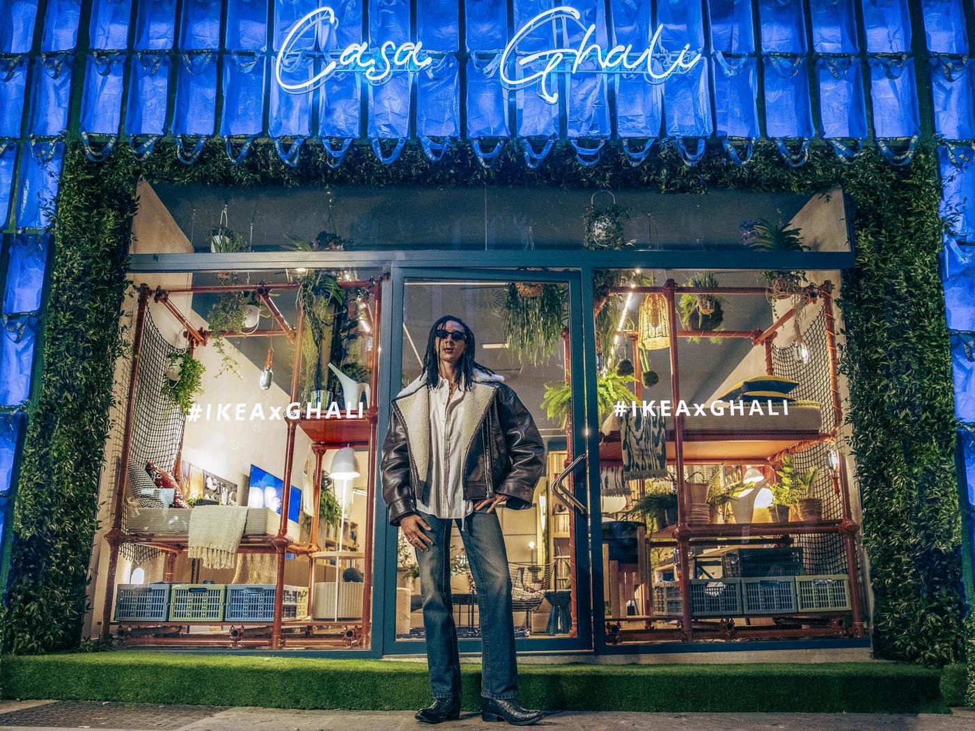 Ghali fuori dalla vetrina di Casa Ghali sembra accogliere gli ospiti a casa sua. Le pareti laterali della casa sono ricoperte di borse blu FRAKTA e in alto c'è la scritta "casa ghali" in neon blu.