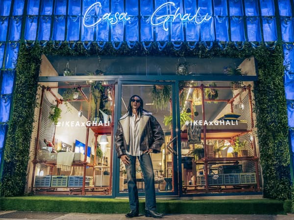 Ghali fuori dalla vetrina di Casa Ghali sembra accogliere gli ospiti a casa sua. Le pareti laterali della casa sono ricoperte di borse blu FRAKTA e in alto c'è la scritta "casa ghali" in neon blu.