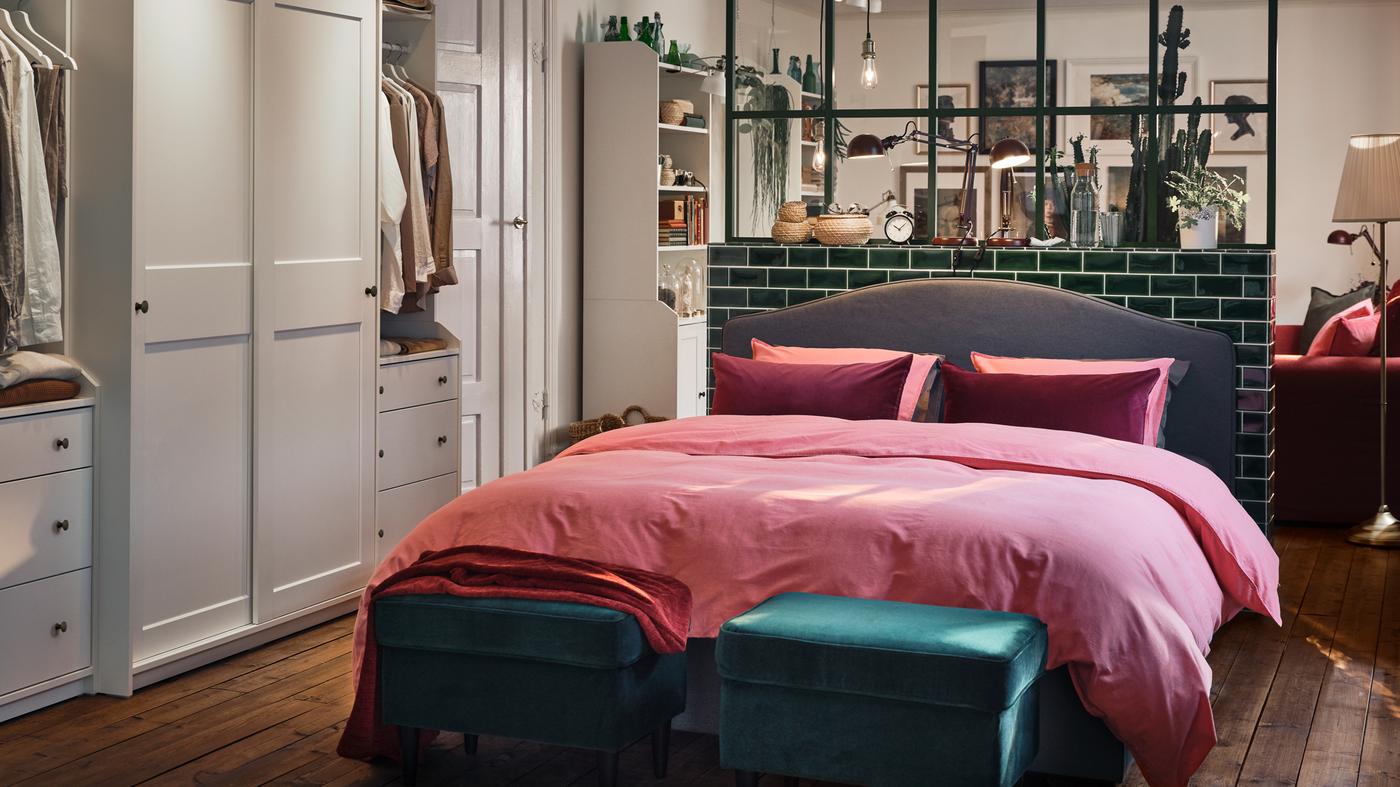 غرفة نوم بها خزانات ملابس بيضاء، وسرير منجد باللون الرمادي، ومنسوجات سرير بلون بني فاتح-أحمر، وزوج من مساند الأقدام بلون أخضر.