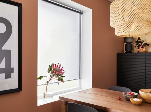 غرفة طعام وردية داكنة مع ستارة لفافة رمادية، ومصباح معلق من الخيزران وطاولة من قشرة خشب البلوط المطلي بالبني.