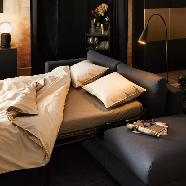 غطاء لحاف KUNGSBLOMMA و اثنين من أغطية المخدات باللون الرمادي والأصفر على كنبة سرير، مع مصابيح وخزانة عرض في الخلف.