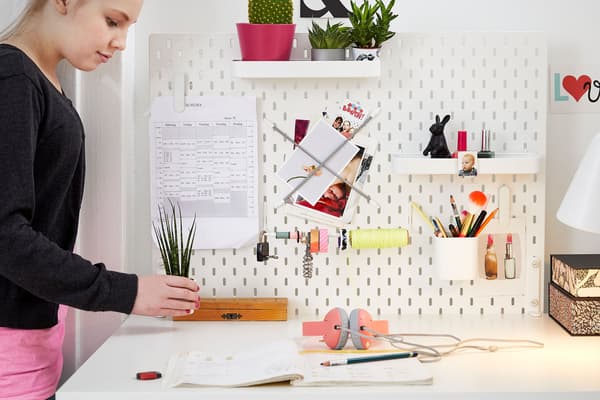 Girl placing small plant onto white desk with a white peg board filled with pens and supplies