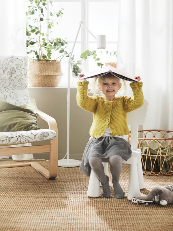 Girl playfully using an open book for a hat sits smiling on a white MAMMUT children’s chair, placed next to a POÄNG armchair.