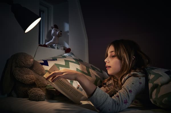 Girl wearing pyjamas reading a book in bed using a small lamp.