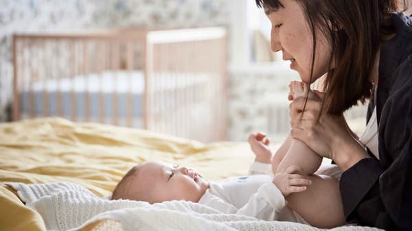 Glad bebis ligger på en vit GULSPARV filt på en säng med sin mamma som håller i hennes fötter och ler mot henne.