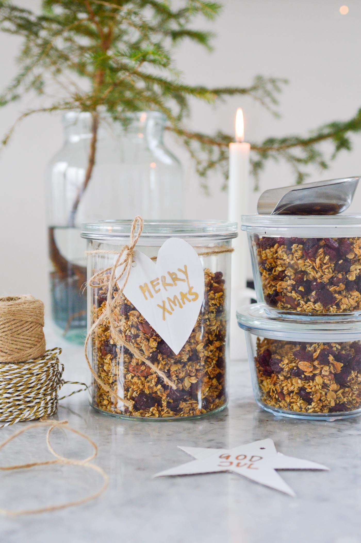Gläser mit Müsli und einem „Merry Xmas”-Etikett stehen auf einer Marmorplatte, umgeben von Schnur, einer weissen Kerze und einem kleinen Baum, was eine festliche, hausgemachte Atmosphäre schafft.