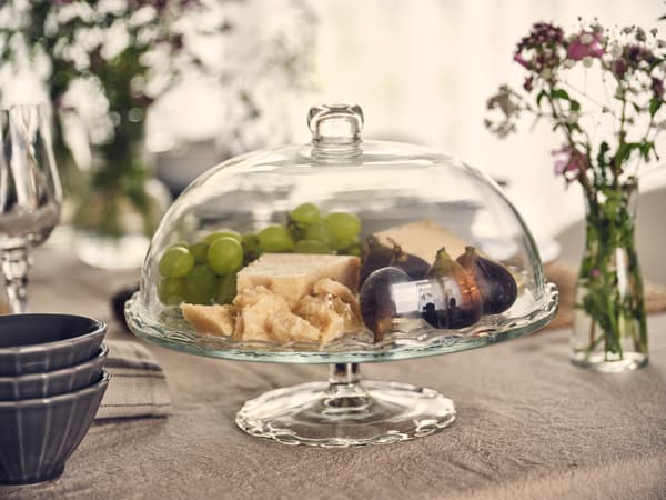 Glass cloche with cheese, grapes and figs on a set table, complemented by flowers and tableware.