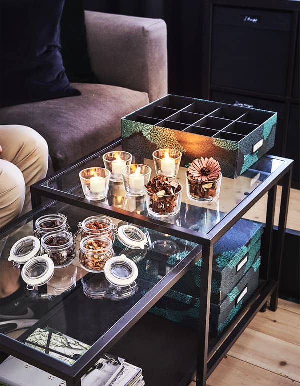 Glass coffee tables decorated with potpourri in jars and candles, with TJENA black and green storage boxes in a living room.