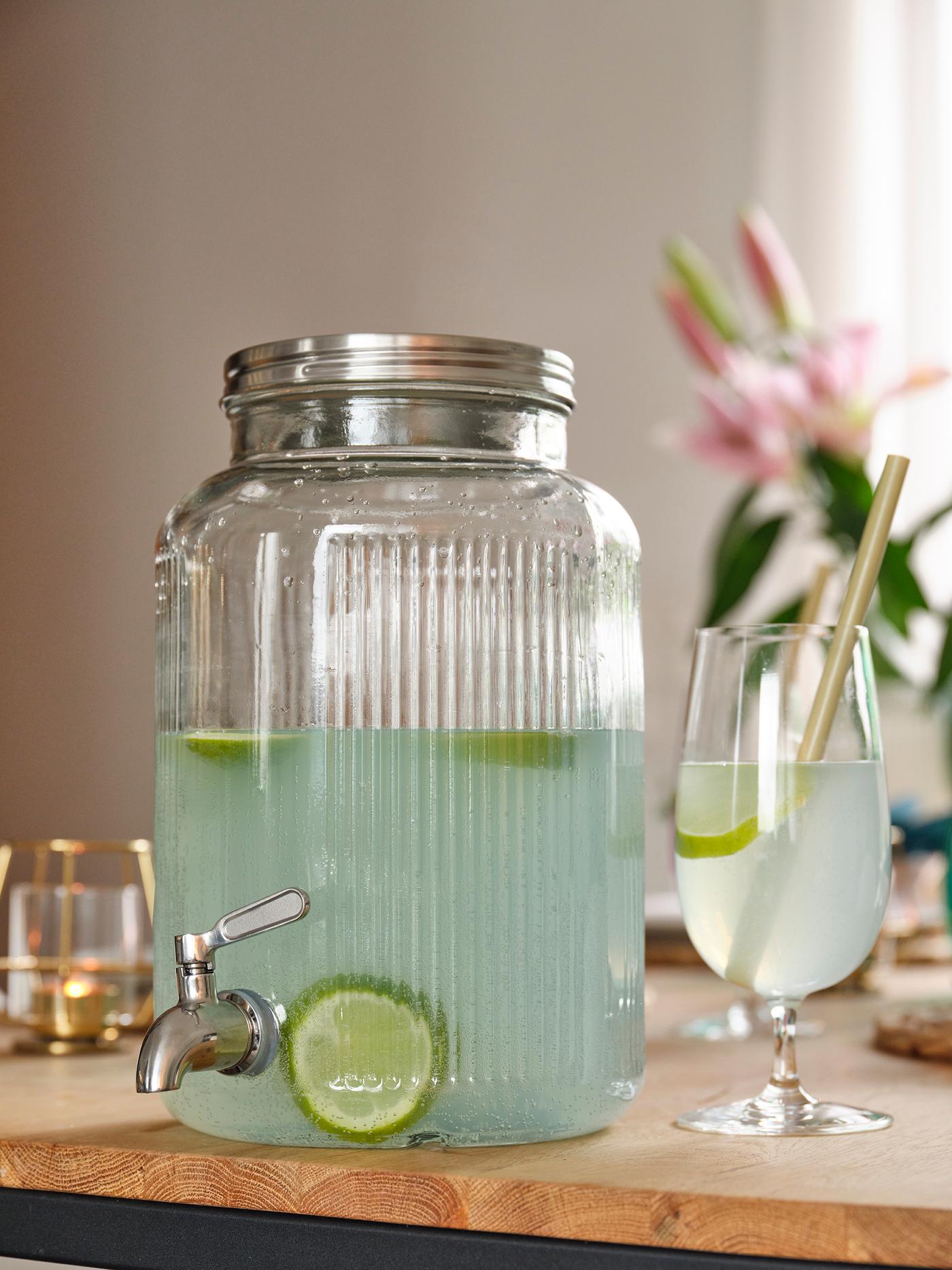Glass drink dispenser with lime water, tap and glass with straw on a summery dining table.