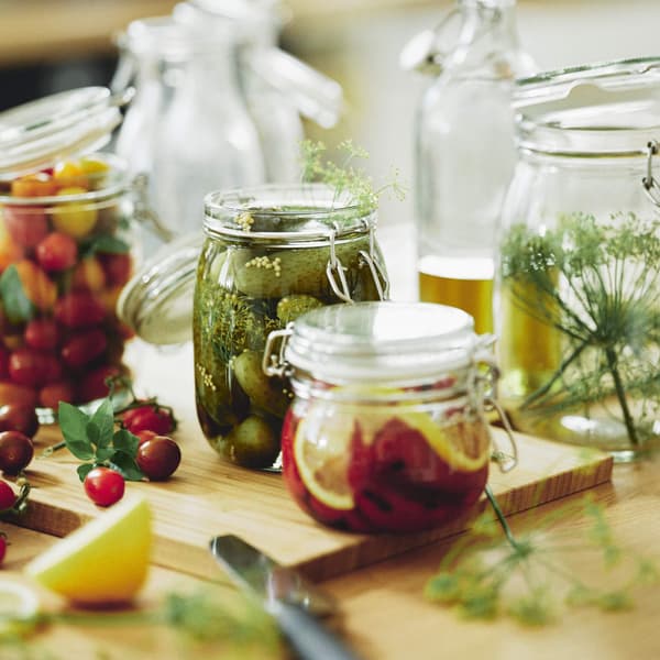 Glass jars of pickled vegetables, fruits, and herbs on a wooden board, surrounded by fresh produce and kitchen items.