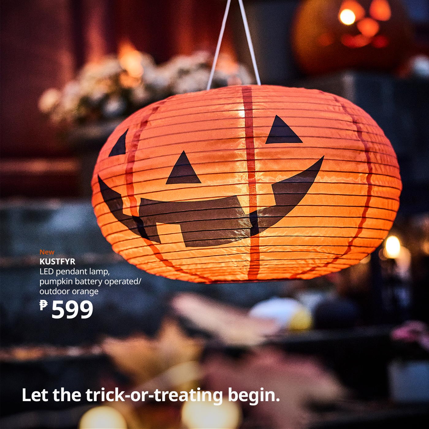 Glowing orange pumpkin-shaped LED pendant lamp with a black jack-o’-lantern face hangs in a cozy shop; Halloween lighting creates a festive mood.