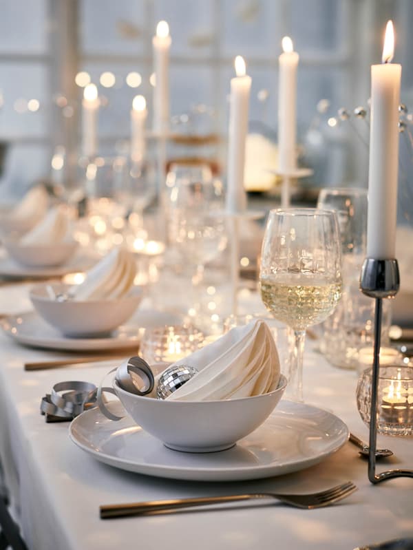 GODMIDDAG tálak és tányérok egy fehér asztalterítővel megterített asztalon, FRÖDJA poharak és SILVERPÄRON gyertyatartók mellett. A gyertyatartókban JUBLA gyertyák égnek. 