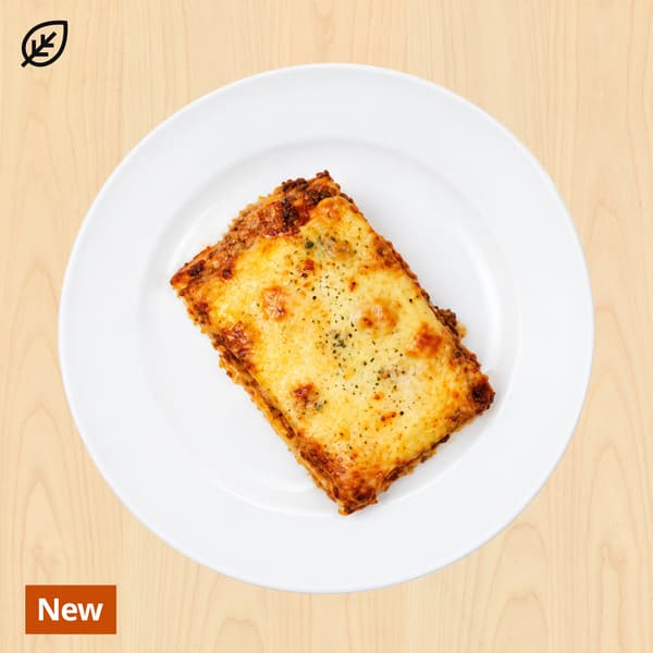 Golden, cheese-topped lasagna slice with browned edges sits on a white plate against a light wooden background; a “New” label marks the dish.