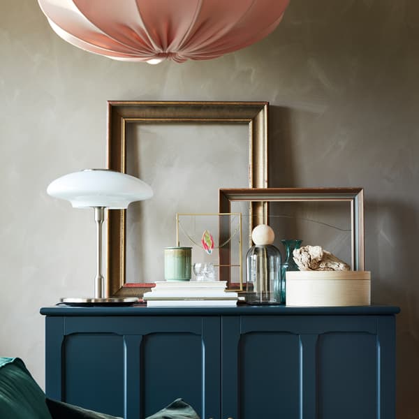 Golden frames, books, decorative items and a table lamp in nickel-plated/opal white glass stand on a dark blue-green cabinet.