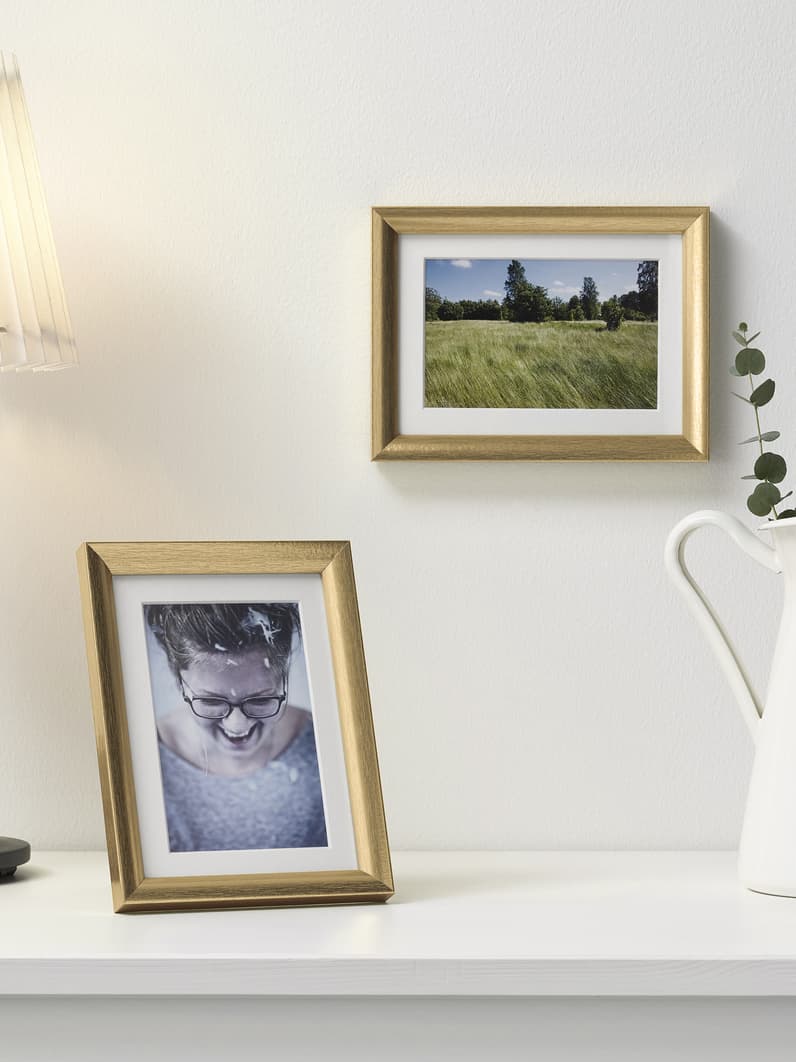 Golden SILVERHÖJDEN photo frames on top of a white storage cabinet next to a table lamp and vase with artificial plants. 